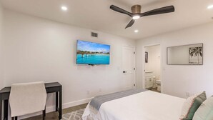 A bedroom with a bed, a desk, a chair, a ceiling fan, a TV, and a bathroom visible through an open door. - 1156 PD - Premium - Luxuriously Renovated Oasis (Siesta Key)