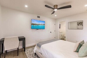 Een slaapkamer met een bed, een bureau, een stoel, een plafondventilator, een televisie en een badkamer, zichtbaar door een geopende deur.