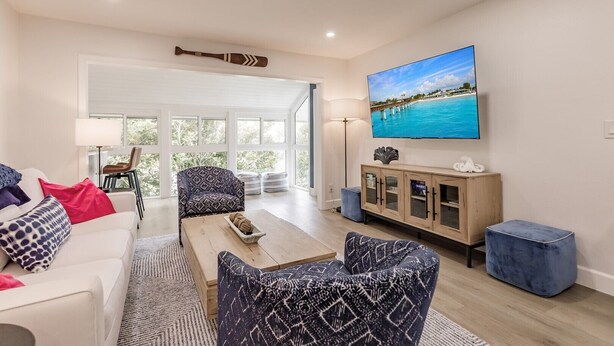 Ein modernes Wohnzimmer mit einem weißen Sofa, gemusterten Sesseln, einem Couchtisch und einem an der Wand befestigten Fernseher, der eine Strandansicht zeigt.