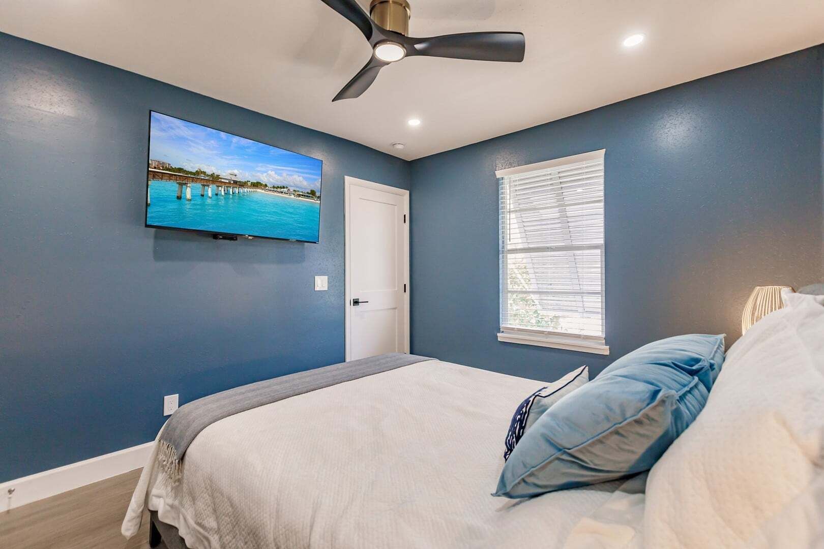 Een slaapkamer met een bed, een plafondventilator, een aan de muur bevestigde televisie en een raam met zonwering.