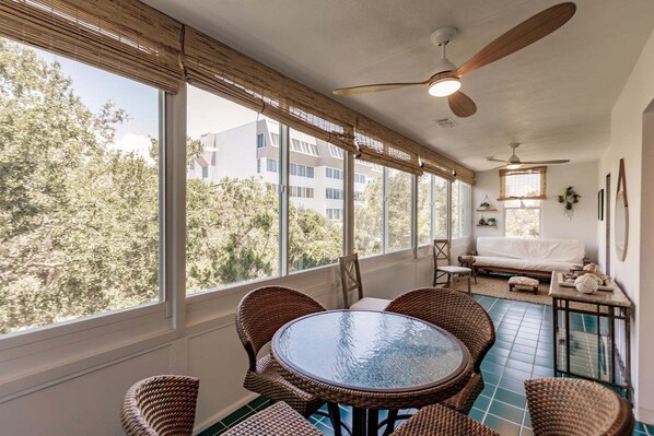 A sunlit room with a round table and wicker chairs, large windows with blinds, a ceiling fan, and a sofa.