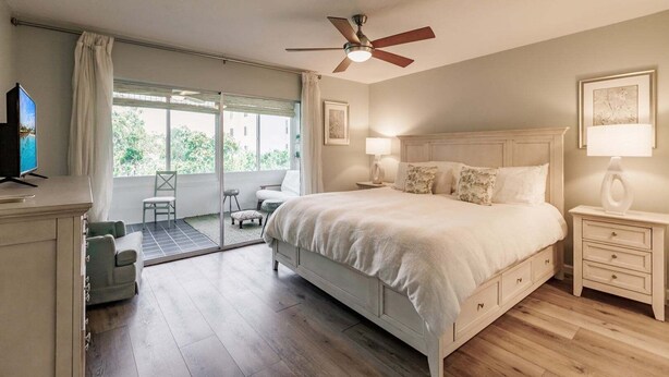 Une chambre à coucher comprenant un lit, des tables de chevet, un ventilateur de plafond, un téléviseur et une porte-fenêtre coulissante donnant sur un espace extérieur aménagé pour se détendre.