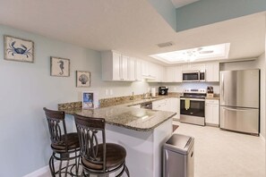 A modern kitchen with white cabinets, a stainless steel refrigerator, a microwave, and a coffee maker. There are two bar stools and a trash can. - 1200-402 - Premium - Experience Serenity Steps from the Water (St. Petersburg)