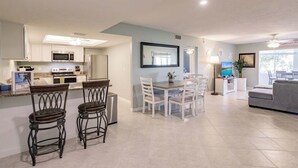 Een moderne keuken met witte kasten, een eetgedeelte met witte stoelen, een grijze bank en een televisie in de woonkamer.