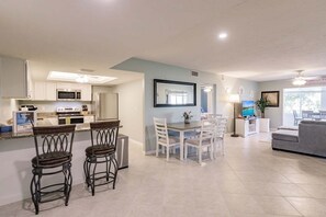 A modern kitchen with white cabinets, a dining area with white chairs, a grey sofa, and a TV in the living room.