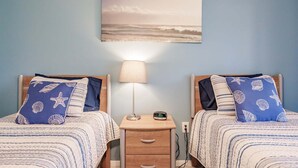 Een slaapkamer met twee bedden, elk voorzien van een kussen met een zeesterren- en schelpenprint, een nachtkastje met een lamp en een aan de muur hangend schilderij van een strandgezicht.