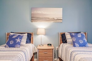 A bedroom with two beds, each with a starfish and shell patterned pillow, a bedside table with a lamp, and a wall-mounted painting of a beach scene.