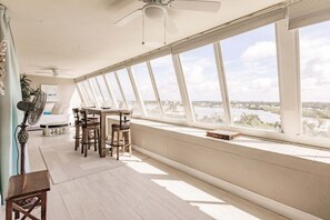 Ein Zimmer mit hoher Decke, einem Esstisch mit Stühlen, einem Ventilator und Blick auf ein Gewässer durch große Fenster.