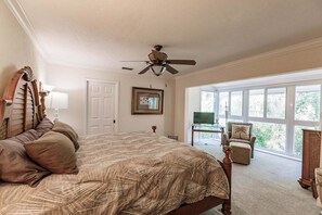 A bedroom with a large bed, a ceiling fan, a television on a stand, and a view of the outdoors through large windows.