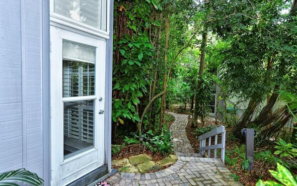 A pathway leading to a white door with a window, surrounded by lush greenery and a stone-paved path. - 6030 EPW - Standard Plus - Cozy Bay Side Garden Paradise (Siesta Key)