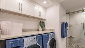 A laundry area with a washing machine and dryer, a countertop with a basket, and a shower area with a glass door. - 6042 - Premium Plus-Ultimate Vacation Haven (Sarasota)
