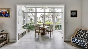 Een ruime kamer met een grote schuifglazen deur naar een balkon met uitzicht op palmbomen en huizen. Er staat een houten eettafel met stoelen en een kamerbank met een patroon.