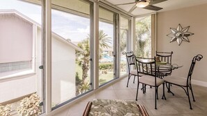 A room with a dining table and chairs, a ceiling fan, and large windows overlooking a landscape with palm trees.