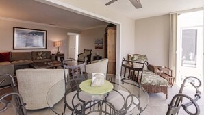 A spacious living area with a glass coffee table, seating arrangements, and a ceiling fan.