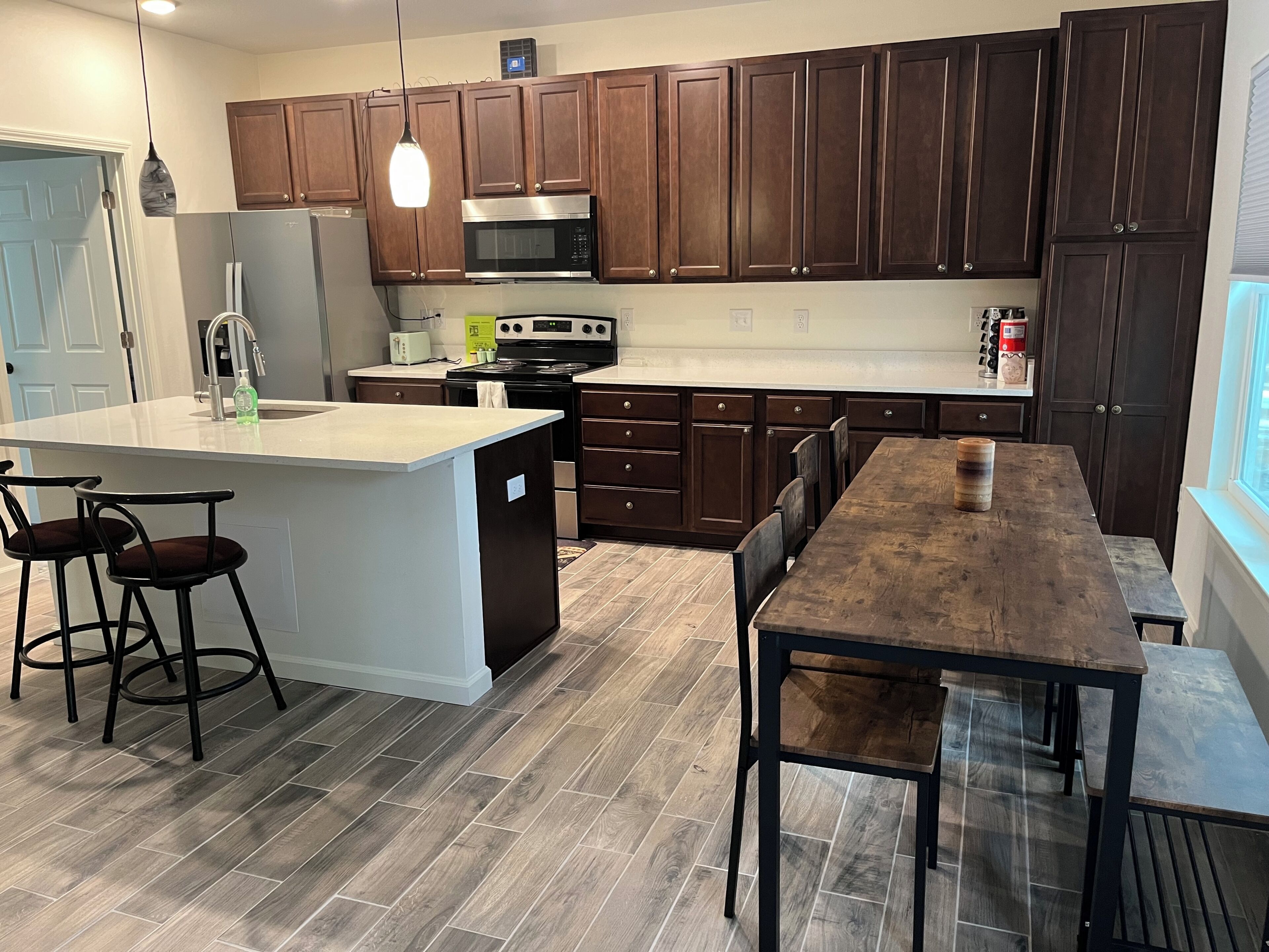 A modern kitchen with dark wooden cabinets, a central island with bar stools, a dining table, and a large window.