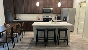 A modern kitchen with dark wooden cabinets, a white countertop, a stainless steel refrigerator, a microwave, a stove, and a sink. There are three black bar stools at the island.