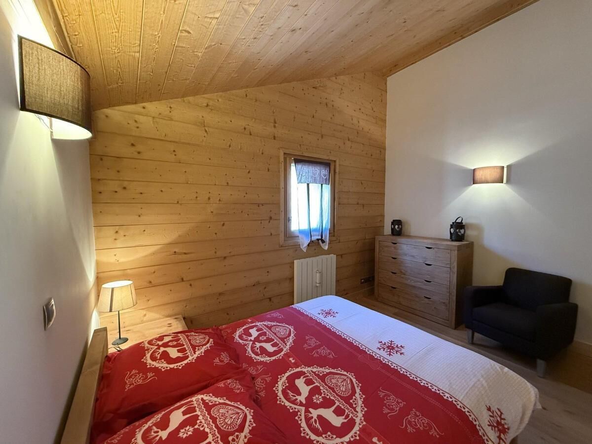 A bedroom with a wooden ceiling, a bed with a red and white patterned bedspread, a dresser, a nightstand with a lamp, and a window with curtains.