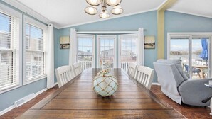 A dining room with a wooden table, white chairs, a chandelier, and large windows with blinds.