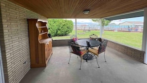 Un patio couvert avec un mur en briques, un meuble en bois, une table ronde avec des chaises, et une vue sur un champ verdoyant et des bâtiments.