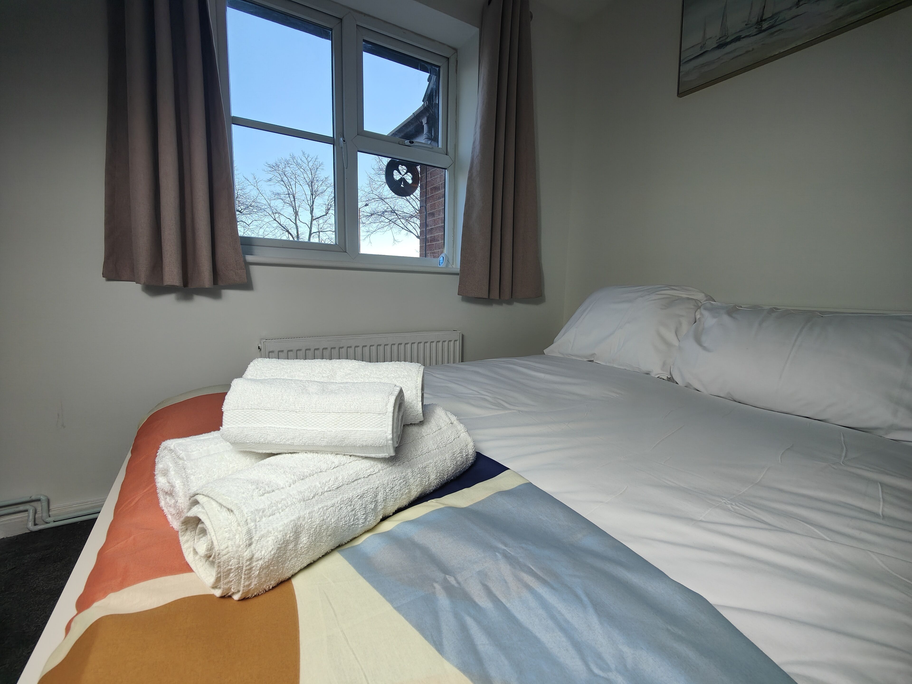 A neatly made bed with white linens and a colorful blanket, a window with curtains, and a radiator.