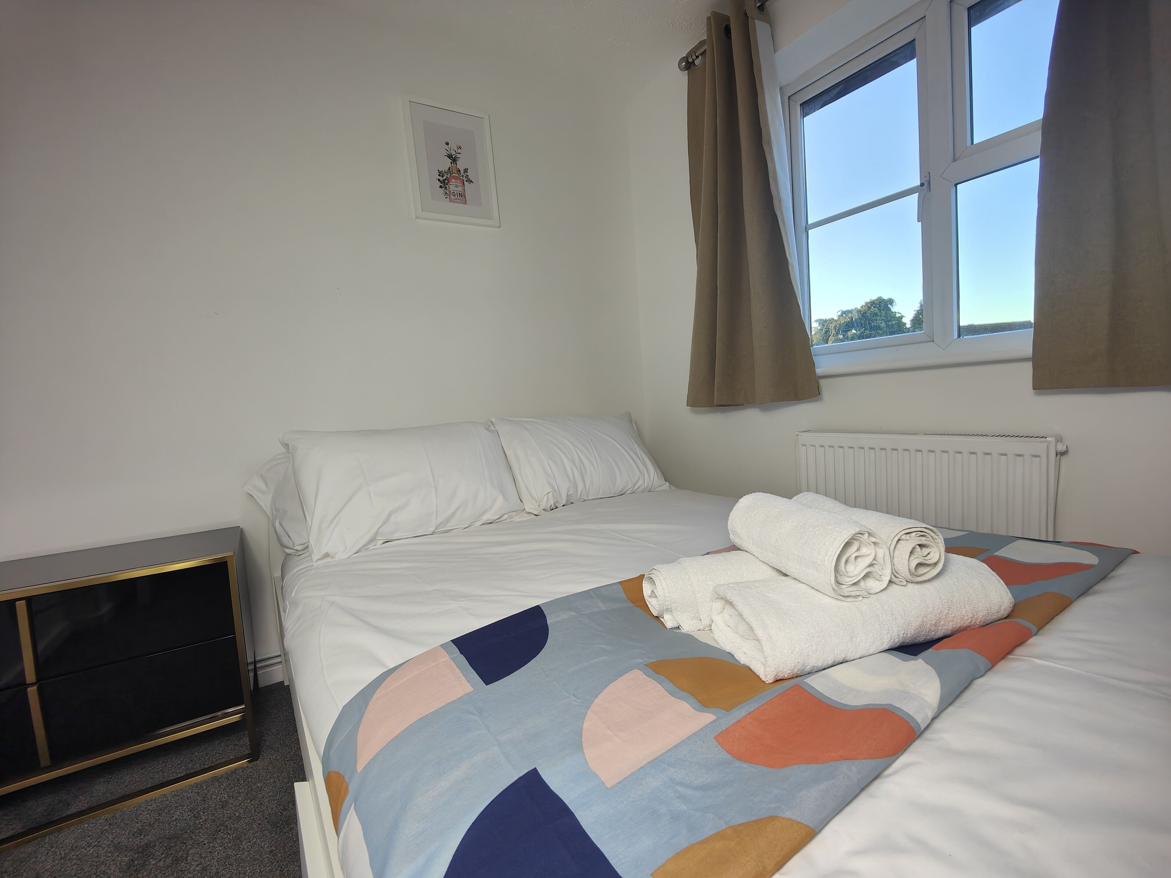 A bedroom with a bed, a radiator, a window with curtains, and a small framed picture on the wall.