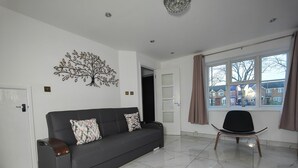 A modern living room with a dark grey sofa, two patterned cushions, a decorative tree wall art, a black chair, and a large window with a view of houses. - 3 Bed House - Sleeps 7 - Garden - Parking - Wifi (Birmingham)