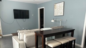 A compact living space with a sofa, a dining table with stools, a TV mounted on the wall, and a door leading to another room. - Marina on the Sakonett (Portsmouth)