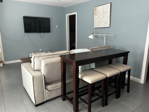 Un espacio habitable compacto con un sofá, una mesa de comedor con taburetes, un televisor montado en la pared y una puerta que da a otra habitación.