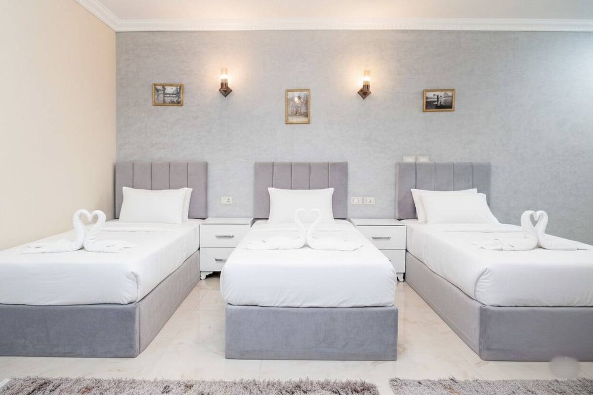 A hotel room with three beds arranged in a row, each with a matching headboard and footboard. There are wall-mounted light fixtures and framed pictures above the beds.