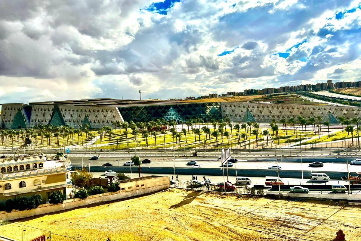 A stadium with a modern design, surrounded by palm trees and a parking area with multiple vehicles.