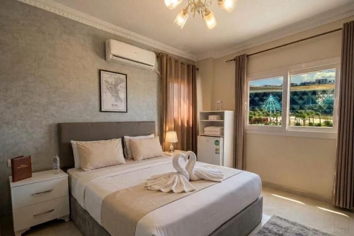 A hotel room with a large bed, a nightstand, a lamp, an air conditioning unit, a window with a view, and a framed picture on the wall.