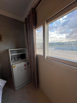 Ett hotellrum med en minibar, ett fönster med utsikt över en parkeringsplats och avlägsna byggnader samt en gardin.