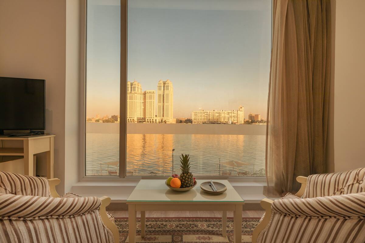 A room with a large window overlooking a body of water and cityscape, a small table with a fruit bowl, and striped armchairs.
