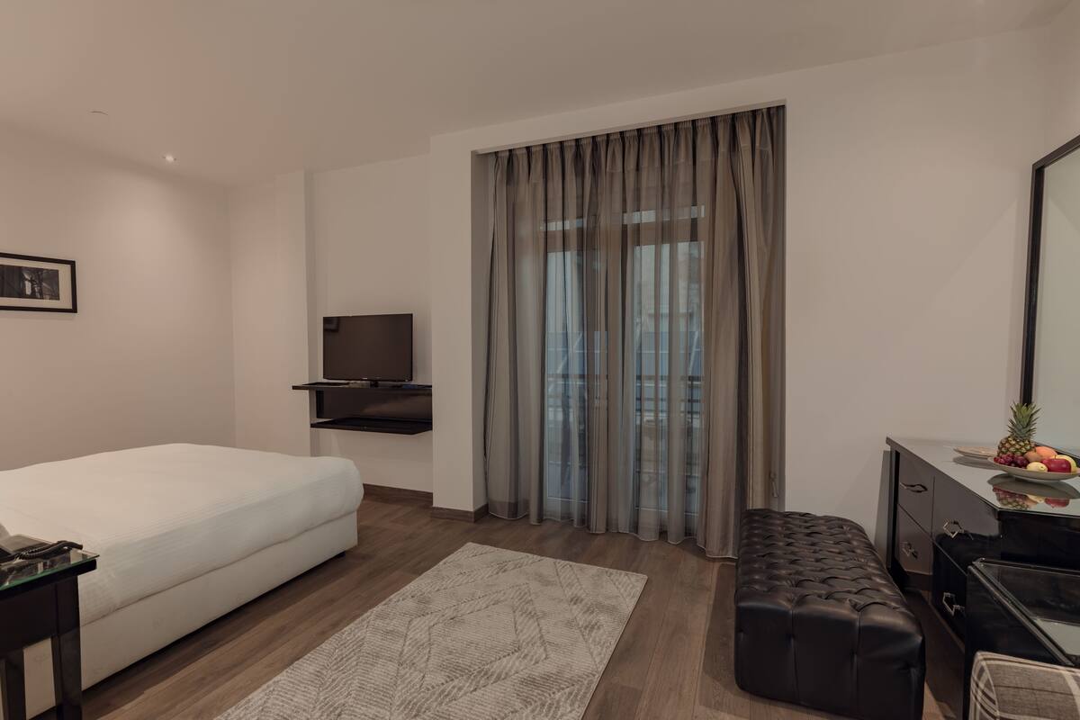 A modern hotel room with a bed, a TV, a desk, a bench, and a large window with curtains.