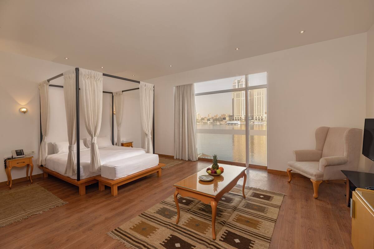 A hotel room with a four-poster bed, a sitting area with a table, a TV, and a large window with a city view.