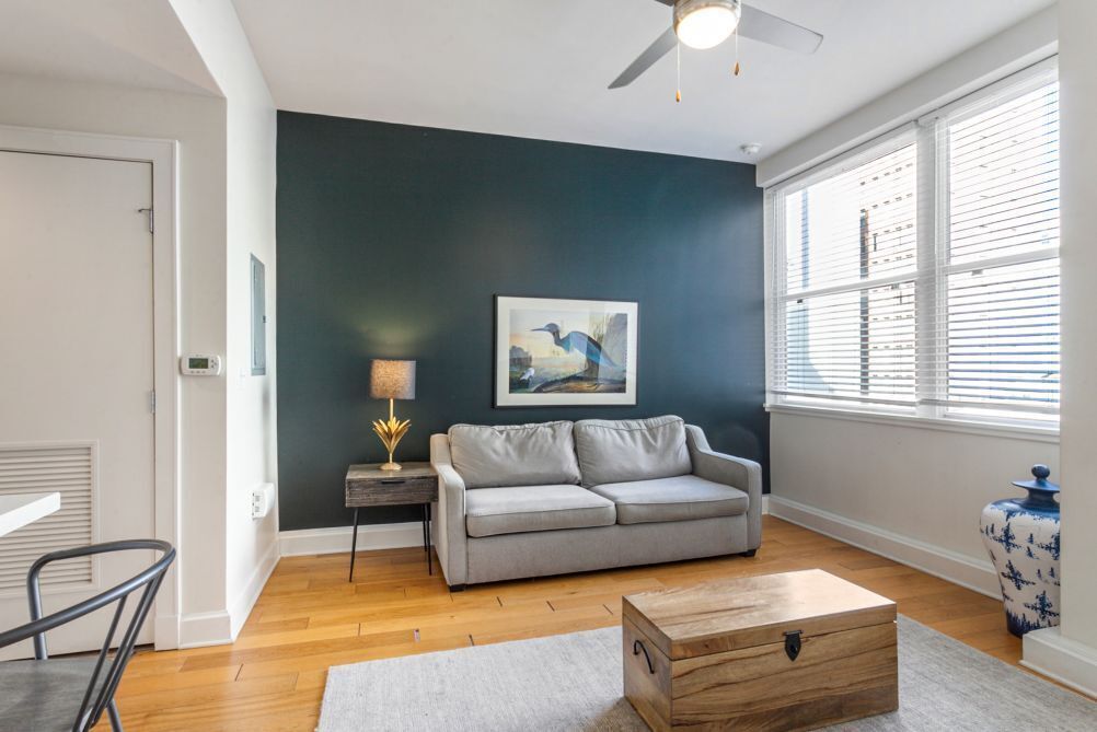 A living room with a grey sofa, a wooden coffee table, a painting on the wall, a lamp, and a window with blinds.