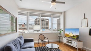Un salon moderne avec un canapé gris, une table basse ronde, un téléviseur posé sur un meuble blanc et de grandes fenêtres équipées de stores.