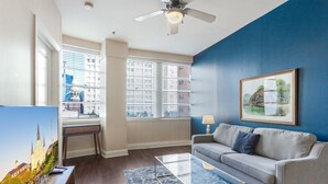 A living room with a sofa, a glass coffee table, a painting, and a desk with a lamp. - Contemporary Comfort: 1BR Condo with Poolside Serenity (New Orleans)