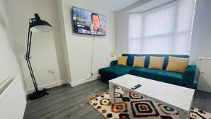 A living room with a teal sofa, a white coffee table, a colorful rug, a black floor lamp, and a TV displaying 'The Terminal'. - Remarkable 4-bed House in Bootle, Liverpool (Bootle)