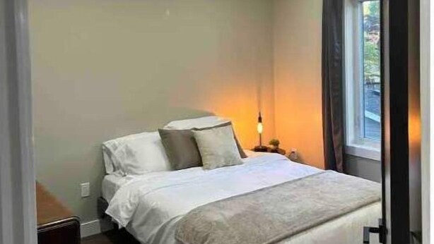 A bedroom with a bed, bedside table, lamp, and a window with curtains. - Gem on Prospect (Kingsville)