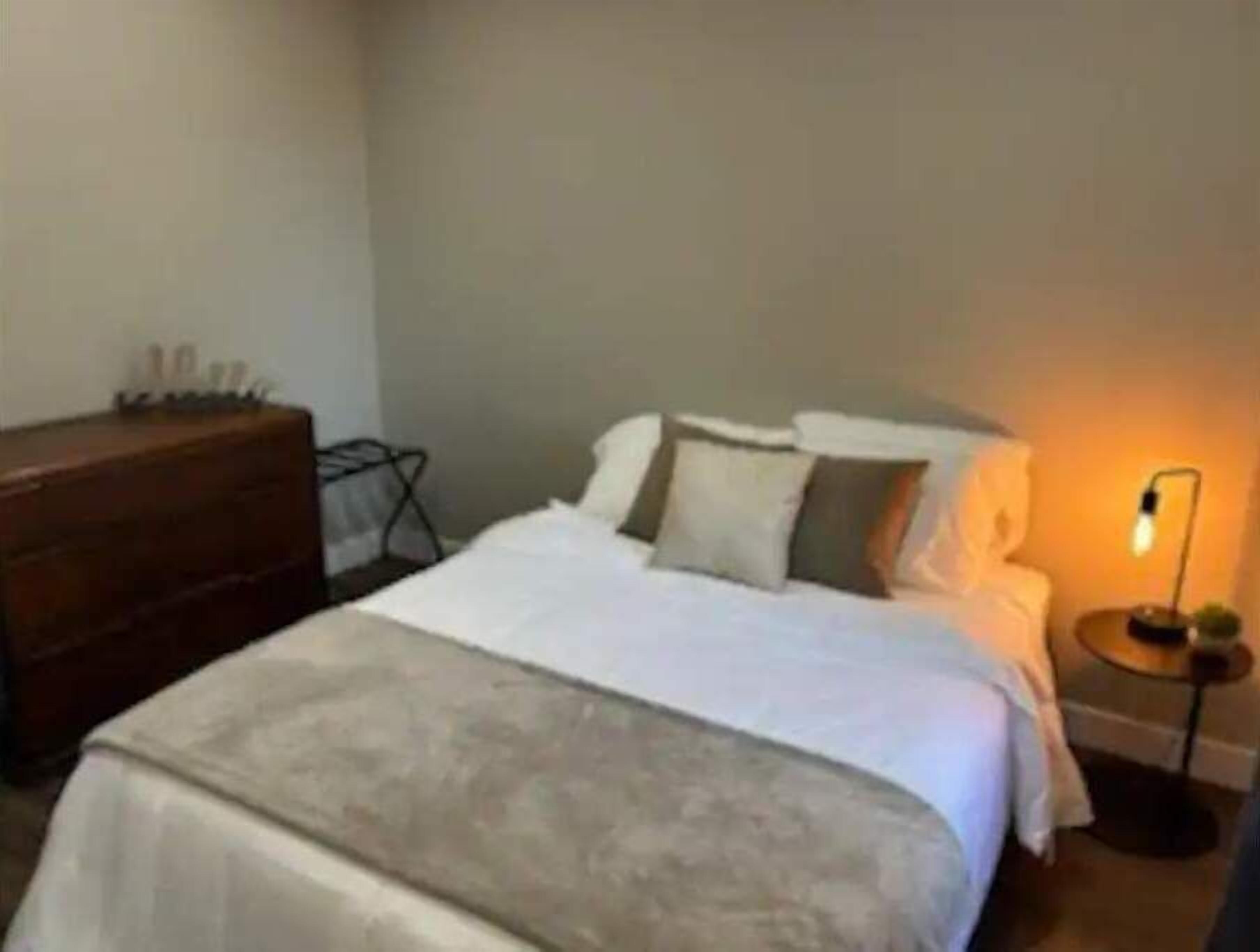 Una cama bien tendida con edredón blanco y almohadas, un aparador de madera y una lámpara de noche sobre una mesita.