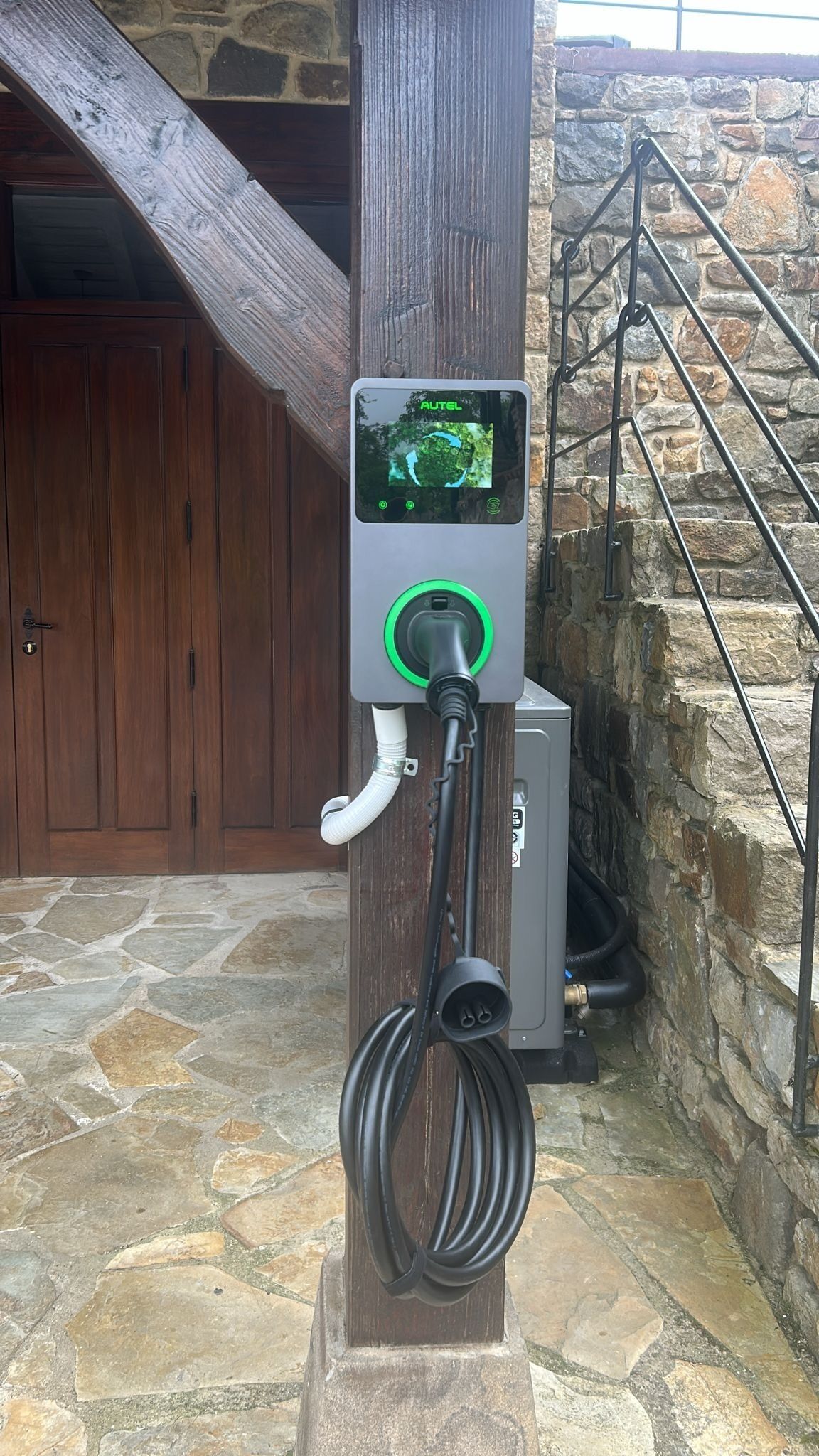 An electric vehicle charging station with a digital display, connected to a building with stone walls and a wooden door.