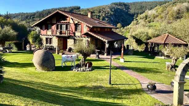 Un chalet à deux étages, doté d’un toit recouvert de tuiles, entouré d’une pelouse bien entretenue et d’un chemin de pierre menant à un belvédère.