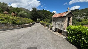 Une route de campagne mène à un bâtiment en pierre, surmonté d’un toit de tuiles, et entouré de verdure et de collines.