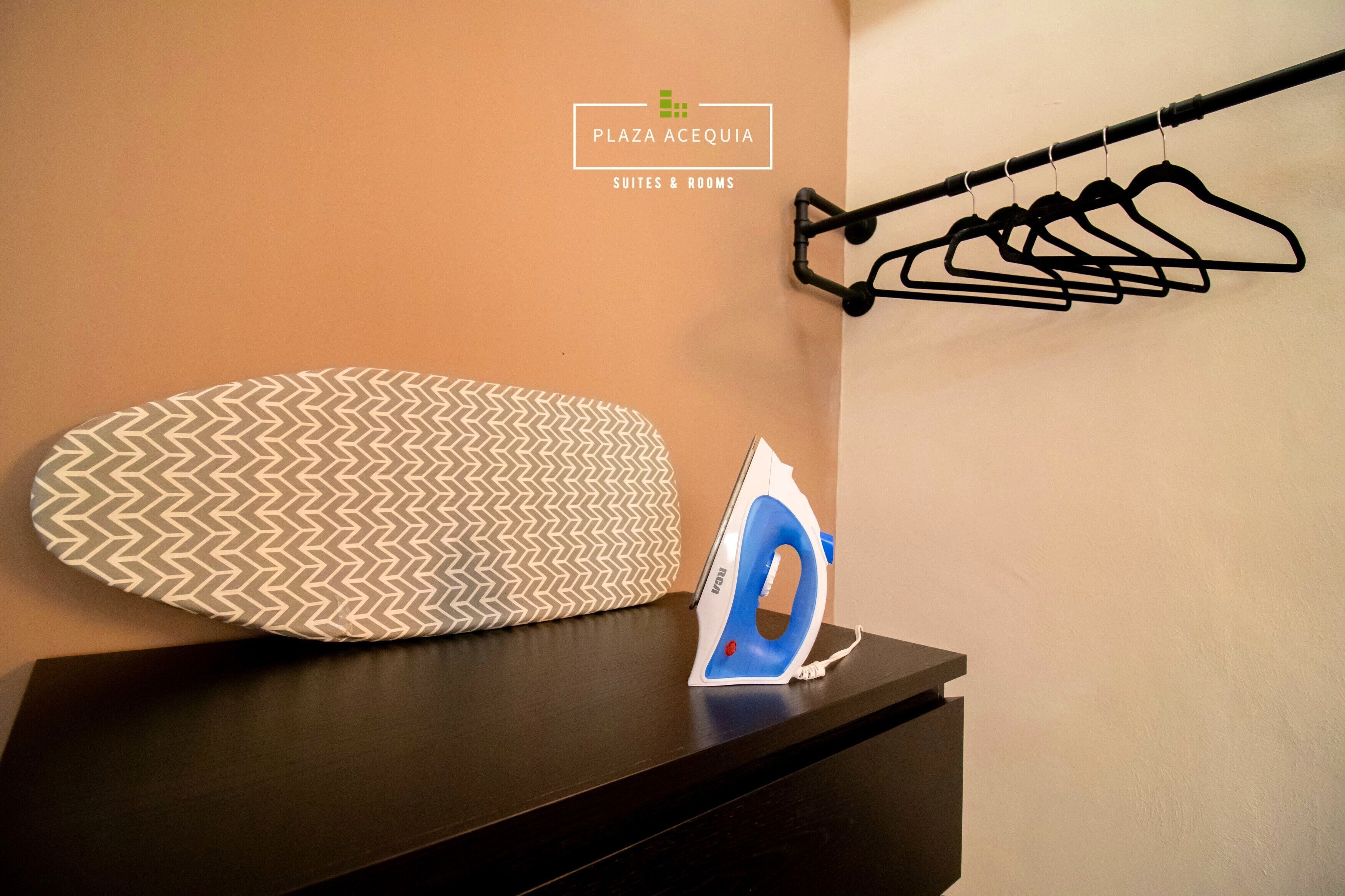 A neatly arranged ironing board with an iron, a hanging clothes rack, and a pillow with a chevron pattern on a dark wooden surface.