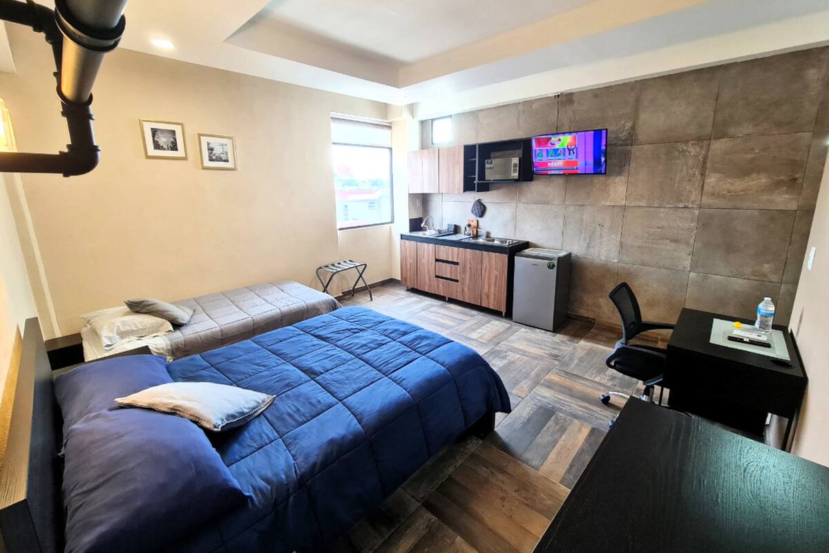 A compact hotel room with a bed, a small kitchenette, a desk, and a TV.