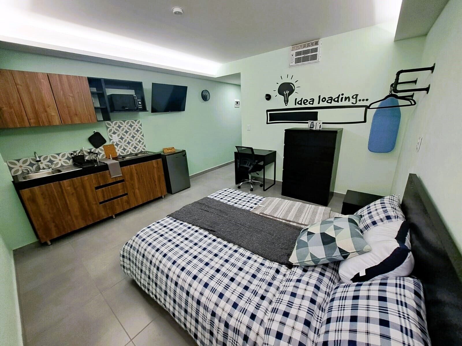Una habitación compacta con cama, un escritorio pequeño, una kitchenette y un televisor montado en la pared.