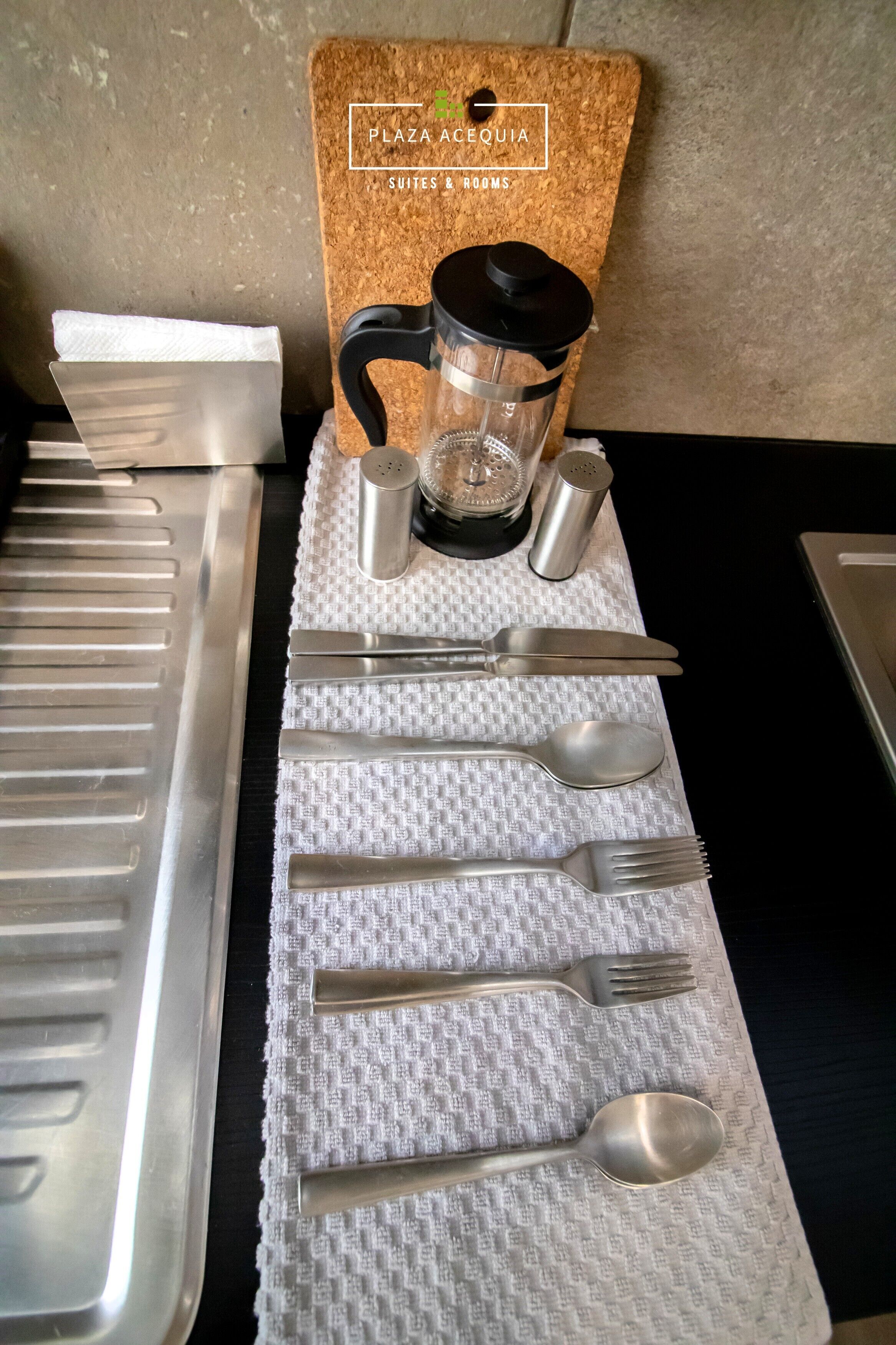 A neatly arranged table with cutlery and a coffee maker on a textured tablecloth, with a cork board and a sign indicating 'PLAZA ACEQUIA SUITES 2 & ROOMS'.