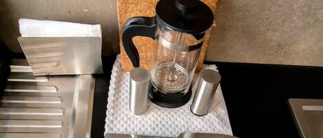 A neatly arranged table with cutlery and a coffee maker on a textured tablecloth, with a cork board and a sign indicating 'PLAZA ACEQUIA SUITES 2 & ROOMS'.