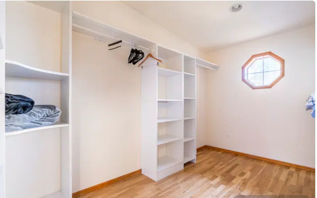 A room with a wooden floor, white walls, a closet with open shelves, and a window with a hexagonal design.